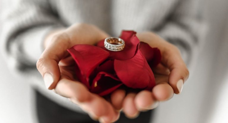ženské ruky držiace lupene červených ruží na ktorých je uložený prsteň