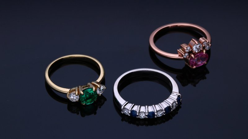 tri prstene z žltého, bieleho a ružového zlata s drahými kameňmi na tmavom pozadí