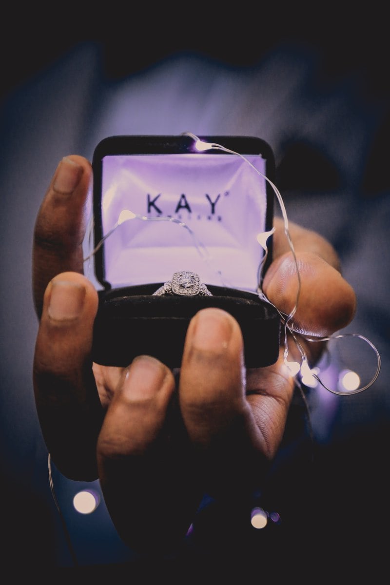 mužská ruka drží zásnubný prsteň v ozdobnej krabičke omotanej svetielkami