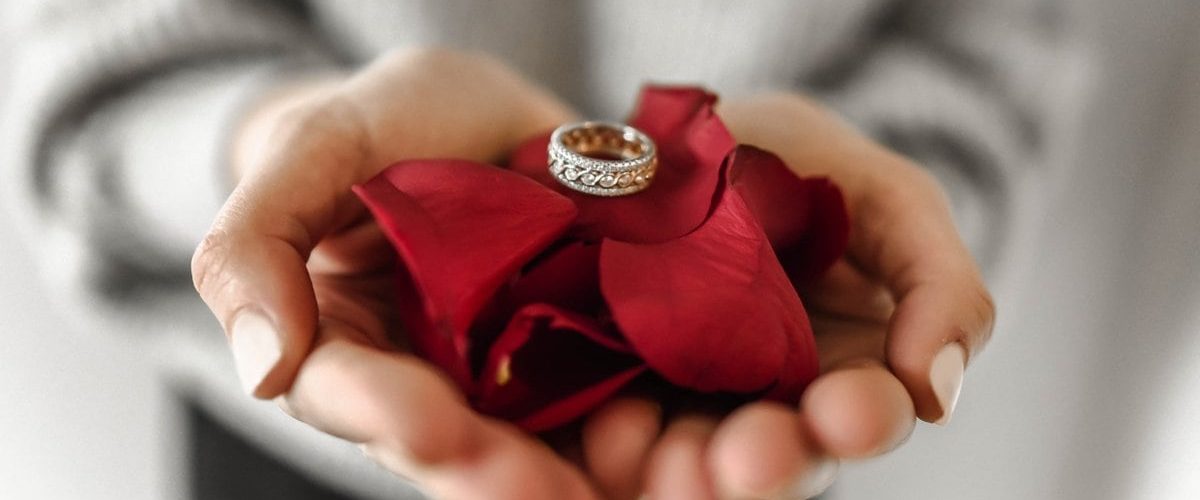 ženské ruky držiace lupene červených ruží na ktorých je uložený prsteň