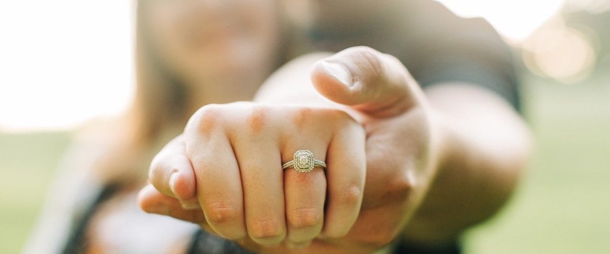muž objíma žena zozadu držiac jej ruku s detailom na diamantový zásnubný prsteň